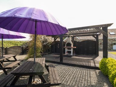 An outdoor seating area with wooden picnic tables and purple umbrellas next to a wooden pergola with a barbecue grill at Bodrual Cottage in Rhosbodrual near Caernarfon