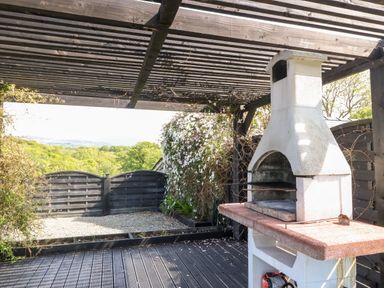 An outdoor patio with a wood pergola a white barbecue grill and a black wooden gate overlooking trees at Bodrual Cottage in Rhosbodrual near Caernarfon