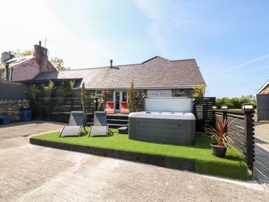 An outdoor area with a hot tub two lounge chairs a stone building and black wooden fences at Bodrual Cottage in Rhosbodrual near Caernarfon