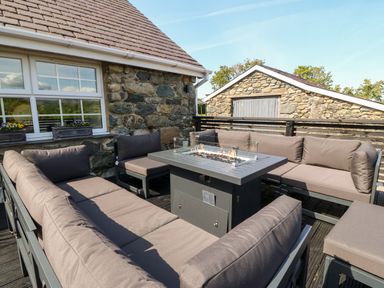 An outdoor seating area with cushioned sofas and a fire pit table on a wooden deck at Bodrual Cottage in Rhosbodrual near Caernarfon