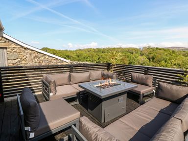 An outdoor seating area with cushioned sofas and a fire pit table on a wooden deck with a view of trees at Bodrual Cottage in Rhosbodrual near Caernarfon