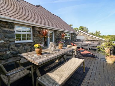 An outdoor patio with a wooden dining table, chairs, benches, potted plants, and lounge chairs at Bodrual Cottage in Rhosbodrual near Caernarfon