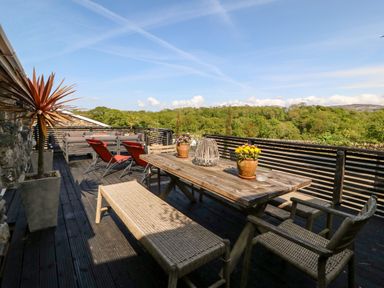 An outdoor wooden deck with a table, bench, chairs, potted plants, lounge chairs, and a seating area surrounded by trees at Bodrual Cottage in Rhosbodrual near Caernarfon
