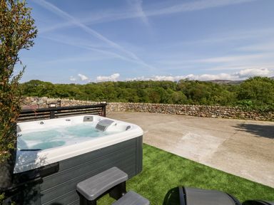 An outdoor hot tub on artificial grass near a stone wall with trees and hills in the background at Bodrual Cottage in Rhosbodrual near Caernarfon