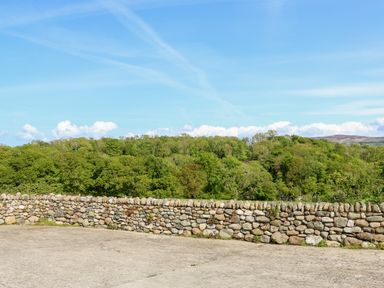 A stone wall with green trees and hills under a blue sky at Bodrual Cottage in Rhosbodrual near Caernarfon