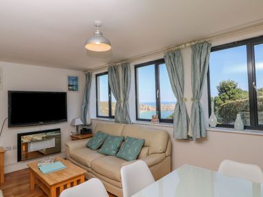 A living room with a beige sofa and blue cushions near windows with blue curtains and a wall mounted TV at Beachcomber in Porthcurno near Sennen