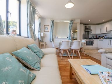 A living room with a cream sofa and turquoise cushions a dining table with white chairs and an open kitchen in the background at Beachcomber in Porthcurno near Sennen
