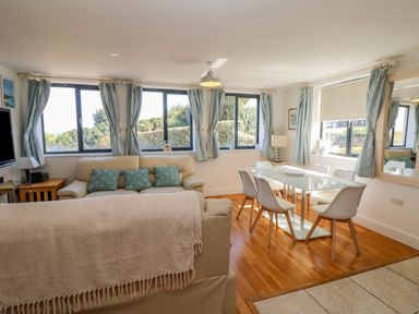 A dining area with a glass table and six white chairs next to a living room with two cream sofas and blue cushions at Beachcomber in Porthcurno near Sennen