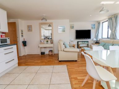 An open living space with a dining table and chairs a beige sofa a TV on the wall a mirror and a kitchen area at Beachcomber in Porthcurno near Sennen