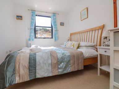 A bedroom with a wooden bed frame a patchwork bedspread cushions spelling sea blue curtains and a window at Beachcomber in Porthcurno near Sennen