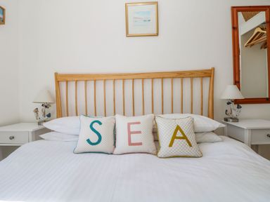 A bedroom with a wooden bed frame white bedding three pillows spelling SEA two white nightstands with lamps a framed picture on the wall and a mirror showing hangers at Beachcomber in Porthcurno near Sennen
