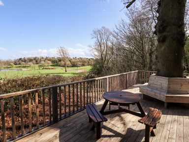 A wooden deck with a round picnic table and benches and a bench built around a tree overlooking a green field at iLodge Ultra in Kenwick near Louth