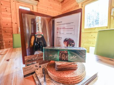 A wooden table with a bottle of Asti, After Eight chocolates, a welcome folder, coasters, a pen, and discount cards inside a wooden cabin at iLodge 73 Kenwick near Louth