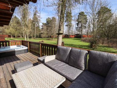 A wooden deck with a hot tub and grey cushioned outdoor seating overlooking a grassy area with trees and wooden lodges at iLodge 73 Kenwick near Louth