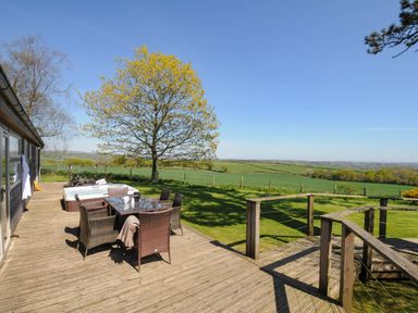 An outdoor wooden deck with a table and chairs next to a grassy field and a tree at 10 Faraway Fields in Dobwalls