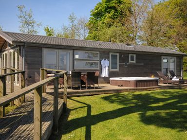 A wooden patio with dining chairs a hot tub and lounge chairs outside a lodge surrounded by trees at 10 Faraway Fields in Dobwalls