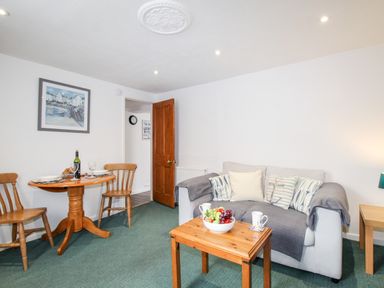 A living room with a sofa and cushions a coffee table with fruit a dining table with chairs and a bottle of wine at Angel Cottage in Weymouth