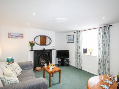 A living room with a sofa, coffee table, TV, fireplace, window with curtains and a small dining table at Angel Cottage in Weymouth