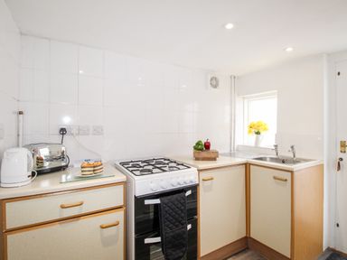 A kitchen with a white cooker oven a kettle toaster a cutting board with vegetables and a sink with flowers on the windowsill at Angel Cottage in Weymouth