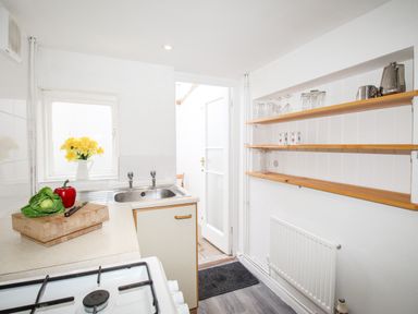 A kitchen with a gas stove, sink, wooden shelves with glasses, and a countertop with vegetables and a knife at Angel Cottage in Weymouth