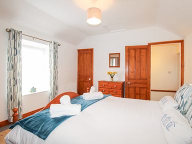 A bedroom with a double bed, wooden doors, chest of drawers with flowers, and a window with patterned curtains at Angel Cottage in Weymouth