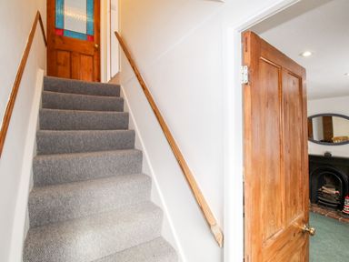 A carpeted staircase with a wooden handrail and a wooden door at the top next to an open wooden door leading to a room with a fireplace at Angel Cottage in Weymouth