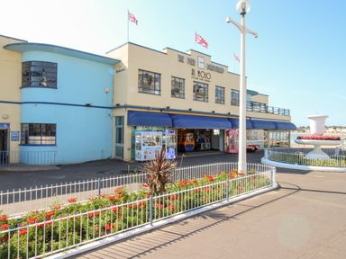 An exterior view of a building with a blue awning and arcade machines under it with a fenced flower bed and a large white planter outside at Angel Cottage in Weymouth