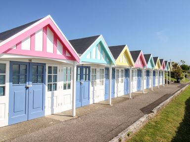 A row of colorful beach huts with blue doors along a paved walkway and grass on the side at Angel Cottage in Weymouth