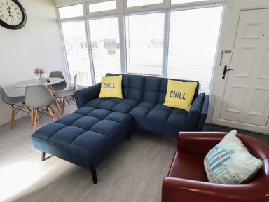 A living room with a blue sectional sofa with yellow pillows a round table with four chairs a red armchair and a wall clock at 23 Cherry Park in Chapel St Leonards
