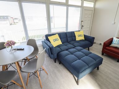 A living room with a blue sectional sofa with yellow chill pillows a round dining table with gray chairs and large windows at 23 Cherry Park in Chapel St Leonards