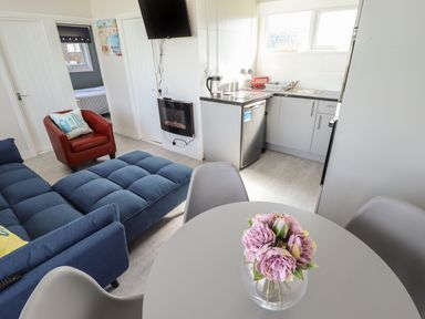 A living area with a round table and flowers a blue sofa a red armchair a kitchen with appliances and a TV mounted on the wall at 23 Cherry Park in Chapel St Leonards