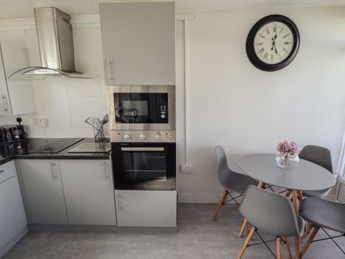 A kitchen with gray cabinets a built-in microwave and oven a round dining table with four gray chairs and a wall clock at 23 Cherry Park Chapel St Leonards