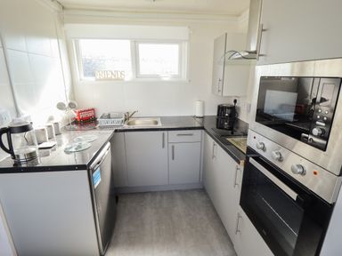 A kitchen with grey cabinets black countertops a dishwasher a kettle a microwave oven and a window with a friends sign at 23 Cherry Park in Chapel St Leonards