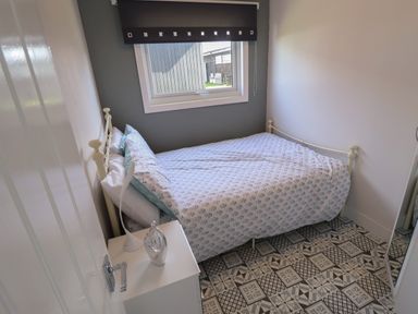 A small bedroom with a patterned tiled floor a white metal-framed single bed with floral bedding a bedside table with a lamp and decorative item and a window with a black window blind at 23 Cherry Park in Chapel St Leonards