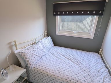 A bedroom with a metal bed frame white bedding a window with a blind and a bedside table with a lamp at 23 Cherry Park in Chapel St Leonards