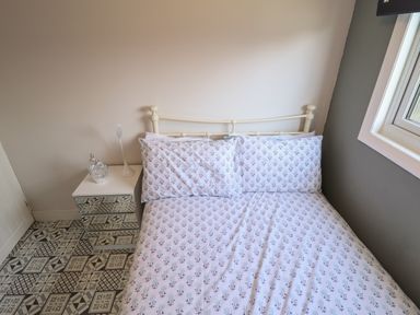 A bedroom with a bed with floral bedding beside a bedside table with a lamp and ornament at 23 Cherry Park in Chapel St Leonards