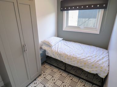 A small bedroom with a single bed covered by white bedding with yellow patterns a gray wardrobe a blue bedside table and a window with a dark blind at 23 Cherry Park in Chapel St Leonards