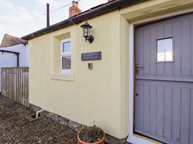 Exterior wall of a cottage with a small window a grey door a light fixture and a potted plant at Garden Cottage in Chathill near Beadnell