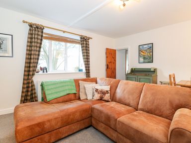 A living room with a brown leather corner sofa green throw and plaid curtains at Garden Cottage in Chathill near Beadnell