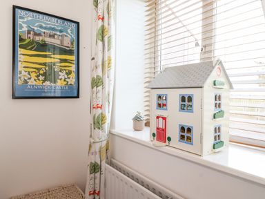 A dollhouse on a windowsill next to a curtain and a framed Northumberland Alnwick Castle poster at Garden Cottage in Chathill near Beadnell