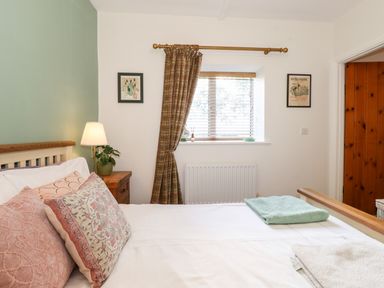 A bedroom with a bed with multiple pillows a bedside table with a lamp and plant a window with blinds and curtains and framed pictures on the wall at Garden Cottage in Chathill near Beadnell