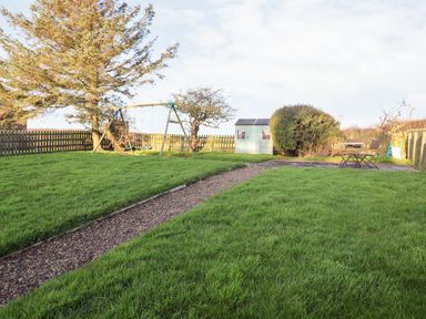 A garden with a gravel path swings a shed a bush and a table with two chairs at Garden Cottage in Chathill near Beadnell