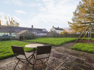 A patio with a small round table and two chairs overlooking a grass yard with a swing set and wooden fence at Garden Cottage in Chathill near Beadnell