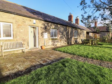 A stone cottage with a wooden bench on a stone patio and a green lawn with outdoor furniture at Garden Cottage in Chathill near Beadnell