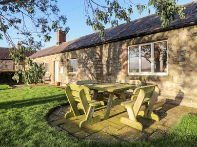 An outdoor wooden table with benches on a stone patio beside a stone house with windows and a door at Garden Cottage in Chathill near Beadnell
