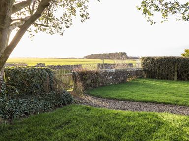 A garden with green grass a gravel path and a wooden gate with stone walls and fields in the background at Garden Cottage in Chathill near Beadnell