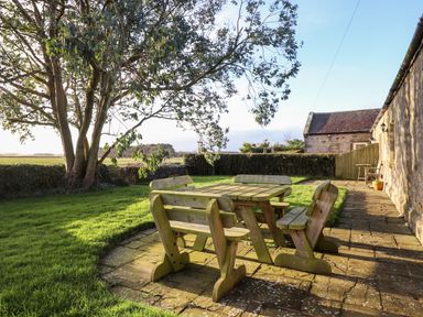 An outdoor patio with a wooden table and four benches next to a stone building and grassy yard at Garden Cottage in Chathill near Beadnell
