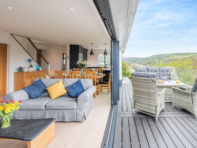 An open living and dining area with a gray sofa and wooden dining table next to an outdoor deck with wicker furniture and a view of green hills at Beach View in Combe Martin