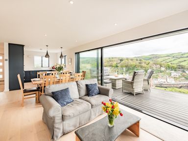 An open plan living and dining area with a sofa and table with flowers next to a sliding door leading to a balcony with outdoor seating and a view of hills at Beach View in Combe Martin