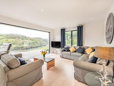 A living room with grey sofas and yellow and blue cushions a coffee table with flowers a TV and large windows with views at Beach View in Combe Martin